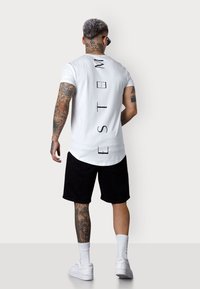 White cotton t-shirt with vertical black text, curved hem, paired with black shorts and white sneakers. Features tattoos on arm and leg.