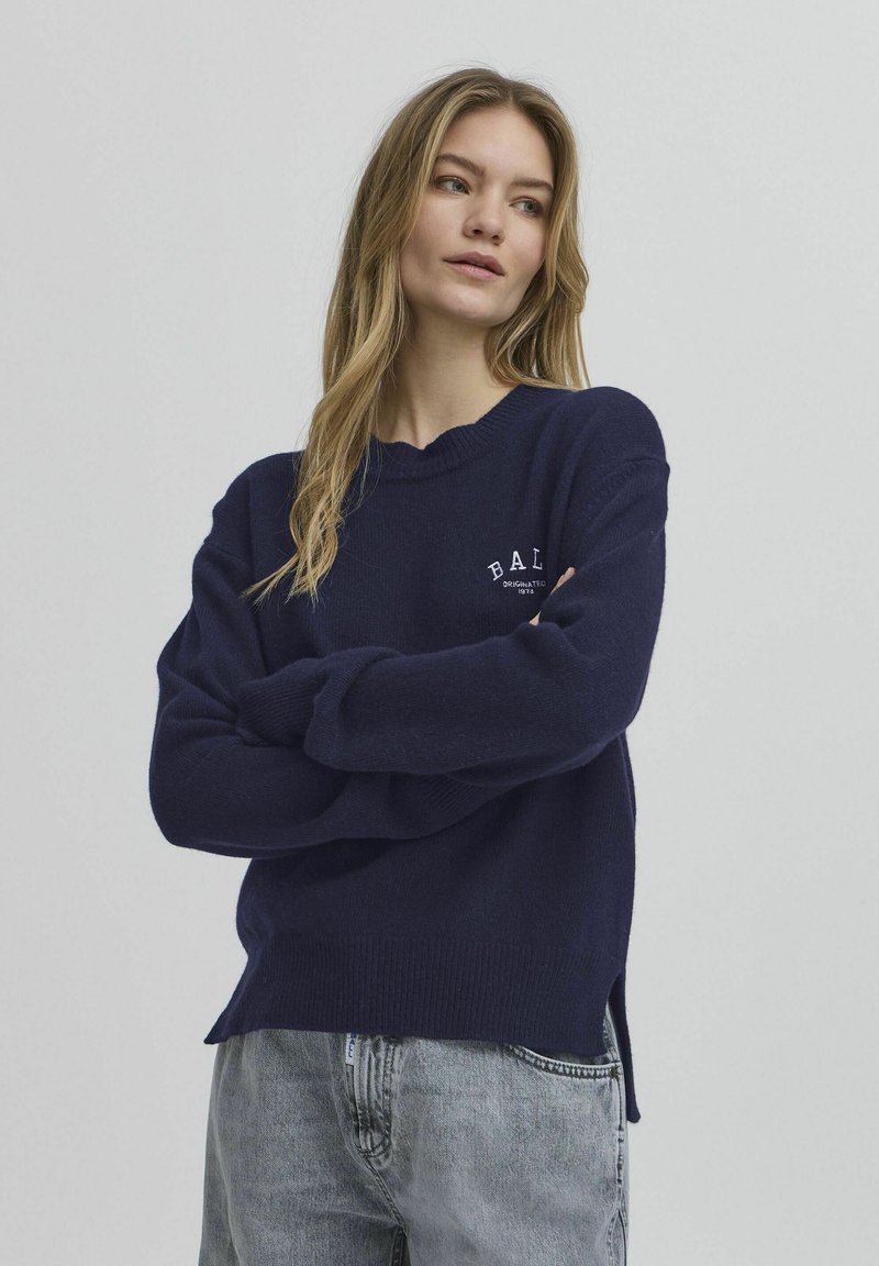 Mørkeblå strikket genser med ribbestrikket hals, løs passform, sidespalter og hvit logo på brystet. Kombinert med grå jeans.