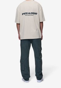 Béžové oversized tričko s logem "PEGADOR" v námořnické modři, spárované s tmavě zelenými cargo kalhotami s kapsami. Vzhled doplňují bílé ploché tenisky.