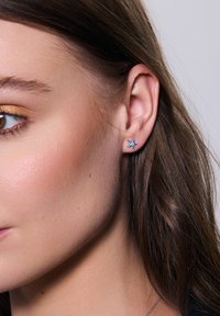 Niebieski kolczyk w kształcie kwiatu, zdobiony licznymi małymi kamieniami. Kolczyk ma wypolerowane metalowe wykończenie oraz bezpieczne zapięcie.