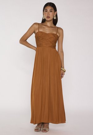 Donna che indossa un abito maxi plissettato color ruggine con spalline sottili, orecchini beige e braccialetti, in piedi davanti a uno sfondo semplice.