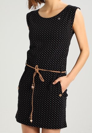 Mouwloze zwarte jurk met witte stippen, bruine gevlochten riem om de taille, voorzakken met knoopdetails, hand in de zak.