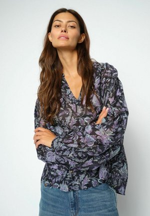 Donna con lunghi capelli castani che indossa una camicetta nera a fiori e jeans blu, in piedi con le braccia incrociate davanti a uno sfondo semplice.