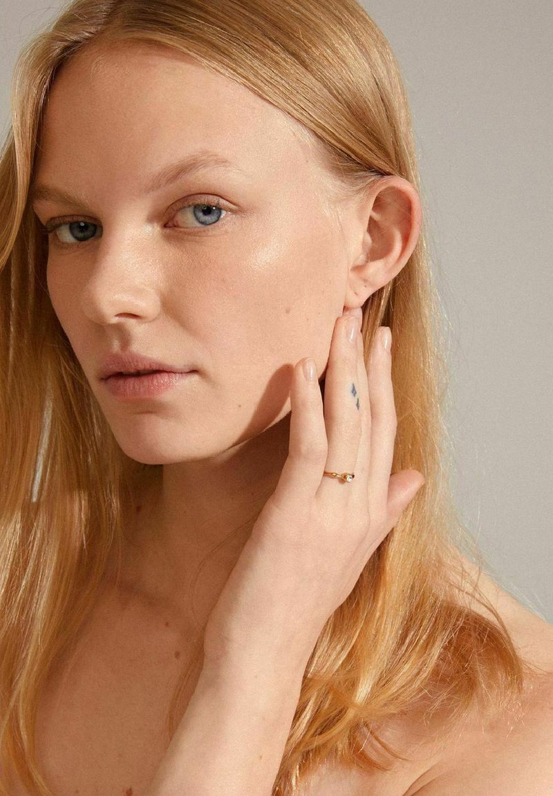 Bague en or avec une petite pierre précieuse transparente, portée à un doigt ; le modèle a de longs cheveux blonds et un teint clair avec un maquillage minimal.