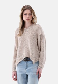 Beige gestrickter Pullover mit V-Ausschnitt, tief sitzenden Schultern und geripptem Saum. Kombiniert mit hellblauen Jeans und einem braunen Gürtel.