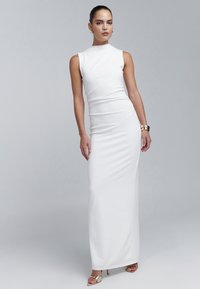 Longue robe blanche sans manches avec un col montant, une silhouette ajustée et une texture lisse. Des bijoux d'accent dorés sont portés aux poignets et aux pieds.