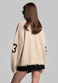 Beiger Oversize-Pullover mit strukturiertem Strick, schwarzer Zahl "23" auf einem Ärmel und fransigen Saumdetails; getragen über einem schwarzen Kleidungsstück.