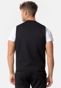 Schwarze Weste aus glattem Stoff, mit einem schlichten Design, hohem Rücken und ohne sichtbare Verschlüsse, getragen über einem weißen Hemd.