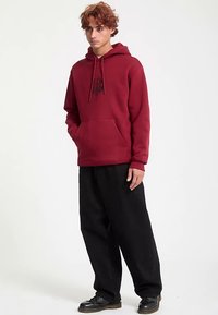 Maroon hoodie with a kangaroo pocket, adjustable drawstrings, and black logo design. Paired with loose-fitting black pants and shoes.