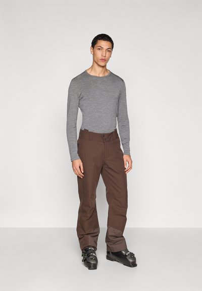 Grå långärmad tröja kombinerad med bruna byxor. Byxorna har en bred midjeresår och är något löst sittande. Svarta skidkängor används.