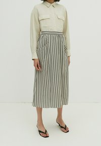 Crème button-up shirt met twee opgestikte zakken, gecombineerd met een zwart-wit gestreepte midi-jurk met zakken en een getailleerde taille. Zwarte sandalen.