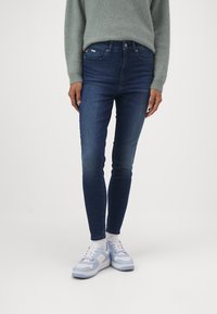 Jeans oscuros ajustados con un acabado desgastado, diseño de cinco bolsillos y una pequeña etiqueta de la marca en la cintura, combinados con zapatillas de deporte de color azul claro.