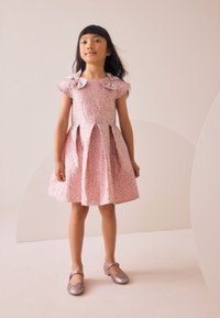 Roosa kleit, millel on tekstuuriga lillemustriga, lühikesed varrukad ja lipsukujuline aksent. Laieneva seelikuga, millel on voldid. Sädelevad roosad kingad.