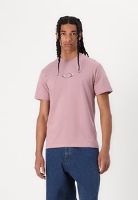 T-shirt de algodão rosa com decote redondo e mangas curtas, apresentando um pequeno logótipo prateado no peito. Usado com jeans azuis.