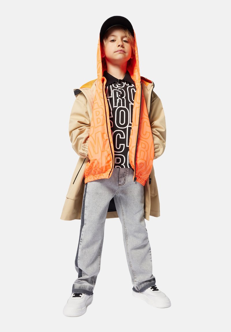 Enfant portant un manteau beige sur un sweat à capuche zippé orange et noir avec un texte géométrique, associé à un jean gris clair et des baskets blanches.