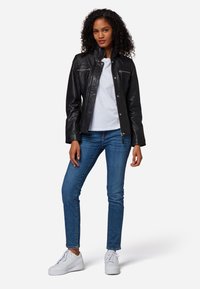 Schwarze Lederjacke mit Reißverschlusstaschen, Druckknopfverschlüssen und hohem Kragen, getragen über einem weißen T-Shirt, kombiniert mit blauen Jeans und weißen Turnschuhen.