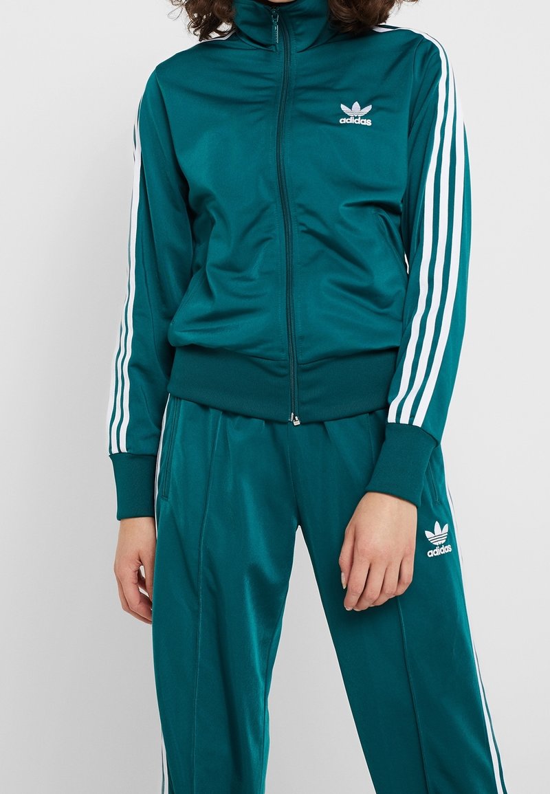 Osoba oblečená v tyrkysovém adidas teplákovém soupravi s bílými pruhy na rukávech a kalhotách, stojící na jednobarevném pozadí.