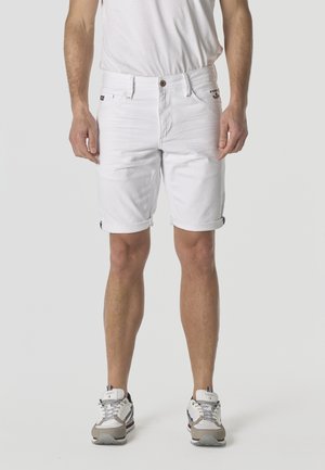 Hombre con pantalones cortos de mezclilla blancos remangados, camiseta blanca y zapatillas blancas con detalles en gris y rojo, de pie contra un fondo liso.