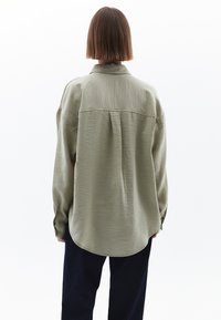 Camicia a maniche lunghe verde oliva chiaro con orlo curvo, tessuto testurizzato, colletto a revers e dettaglio con piega sul retro. Indossata con pantaloni scuri.
