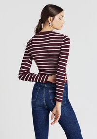 Frau mit braunen Haaren im Pferdeschwanz, die silberne Creolen, ein weinrotes und weiß gestreiftes langärmliges Oberteil und blue Jeans trägt, in seitlicher Pose.