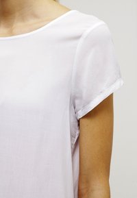 Detailní záběr osoby v jednoduché bílé blůze s krátkým rukávem a výstřihem ve tvaru lodi, na neutrálním pozadí.