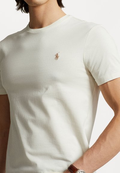 Lys cremfarvet bomulls-t-shirt med rund hals, korte ærmer og et lille kontrastlogo på brystet. Blød tekstur og tætsiddende design.