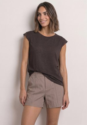 Kvinde med skulderlangt brunt hår iført en mørk ærmeløs top og beige shorts, smilende og kiggende til siden mod en ensfarvet baggrund.