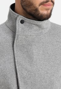 Cappotto grigio in misto lana con colletto alto, linee pulite e chiusura con bottoni laterali. La texture appare morbida con un leggero motivo melangiato.