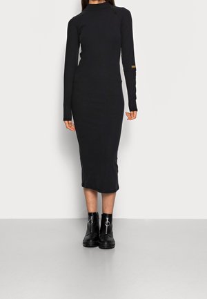 Robe midi noire côtelée avec manches longues, col montant et fente latérale. Associée à des bottines noires à fermeture éclair et à des accessoires circulaires.