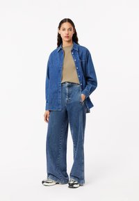 Camisa de denim en azul con un corte relajado, combinada con jeans de tiro alto en azul. La camisa tiene una botonadura en la parte delantera y un bolsillo en el pecho.