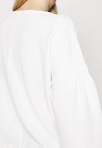 Bílý svetr s texturou a uvolněným střihem. Má nabírané rukávy s elastickými manžetami a jemný žebrovaný vzor u ramenních švů.