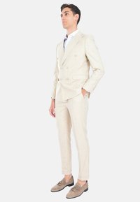 Abito doppiopetto beige chiaro con revers a scollo, abbinato a mocassini in suede marrone chiaro. Pantaloni slim fit, abbinati a una camicia bianca.