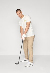 Camiseta blanca de manga corta con gráfico verde, pantalones beiges y zapatillas blancas con acentos negros. Sosteniendo un palo de golf.