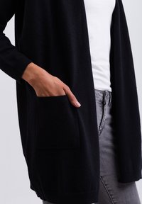 Svart stickad cardigan med öppen framsida, långa ärmar och ribbade ärmslut. Har en patchficka på vänster sida. Bärs över en vit topp.