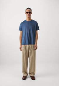 T-shirt di cotone blu con taschino sul petto, abbinato a pantaloni beige a gamba larga e scarpe in pelle marrone. Gli occhiali da sole completano l'outfit.