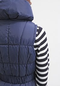 Mörkblå puffer-väst med hög krage, kviltat mönster, samlad midja, bärs över en svart- och vitrandig tröja. Slät, glänsande yta.