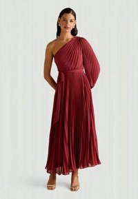 Femme portant une robe bordeaux plissée, jusqu'au sol, à une épaule avec une longue manche et une ceinture assortie, se tenant devant un fond uni.