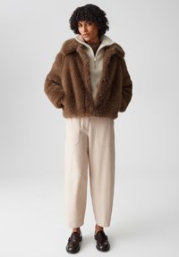 Bruine nepbontjas met een brede kraag, beige gebreide trui eronder, crèmekleurige broek en donkere loafers. Zachte textuur en losse pasvorm.