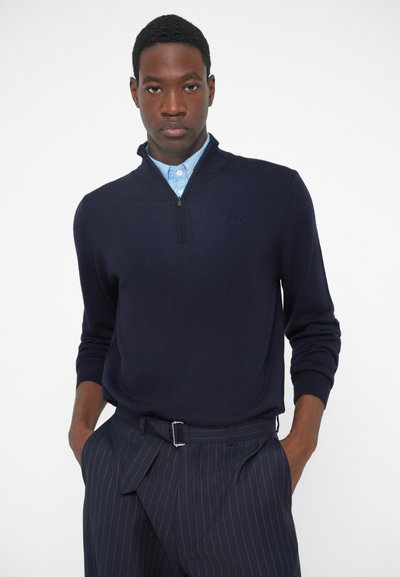 Granatowy sweter zapinany na zamek o teksturowanym wykończeniu, w połączeniu z jasnoniebieską koszulą z kołnierzykiem oraz spodniami w prążki. Wyróżnia się dopasowanym krojem.