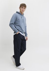 Felpa in cotone azzurro chiaro con cordoncino, indossata sopra una camicia bianca, abbinata a pantaloni blu scuro e sneakers bianche. Vestibilità casual.