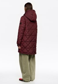 Długa bordowa pikowana kurtka z kapturem, z wzorem w diamenty i ściągaczami przy rękawach, w zestawieniu z luźnymi oliwkowozielonymi spodniami i czarnym obuwiem.