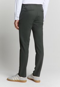 Donkergroene broek met een gladde textuur, elastische tailleband en een achterzak, gecombineerd met lichtgrijze sneakers met witte strepen.