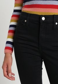 Vaqueros de mezclilla negros de cintura alta con cinco bolsillos, herrajes plateados y un corte ajustado, combinados con un suéter de punto a rayas multicolor.