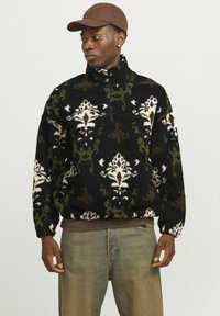 Mikina z fleece v černé barvě s bílými, zelenými a hnědými květinovými vzory; má zipový límec a žebrované cuffs, nošena přes světle modré džíny.