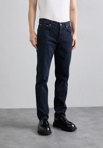 Mørkeblå denimjeans med lige pasform, fem lommer, knaplukning og subtile stikninger, parret med skinnende sorte sko.