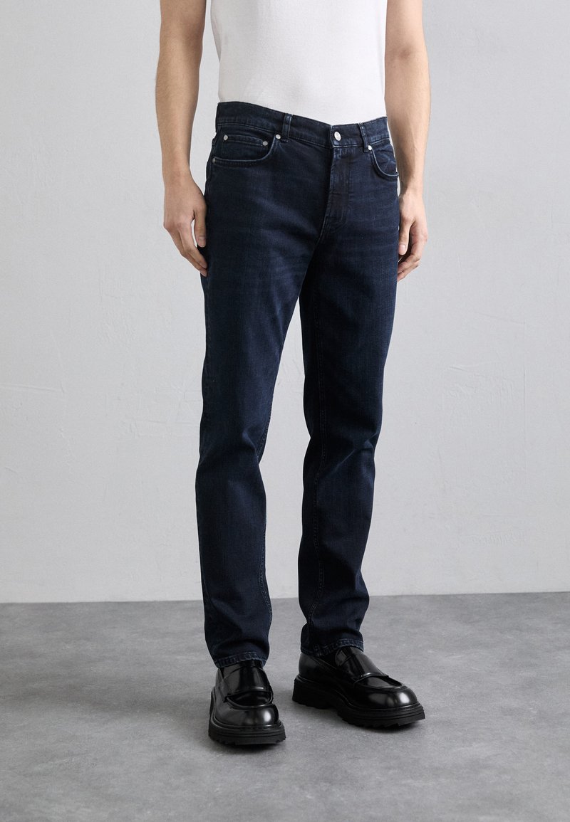 Jeans de mezclilla azul oscuro con un corte recto, cinco bolsillos, cierre de botón y detalles de costura sutiles, combinados con unos zapatos negros brillantes.