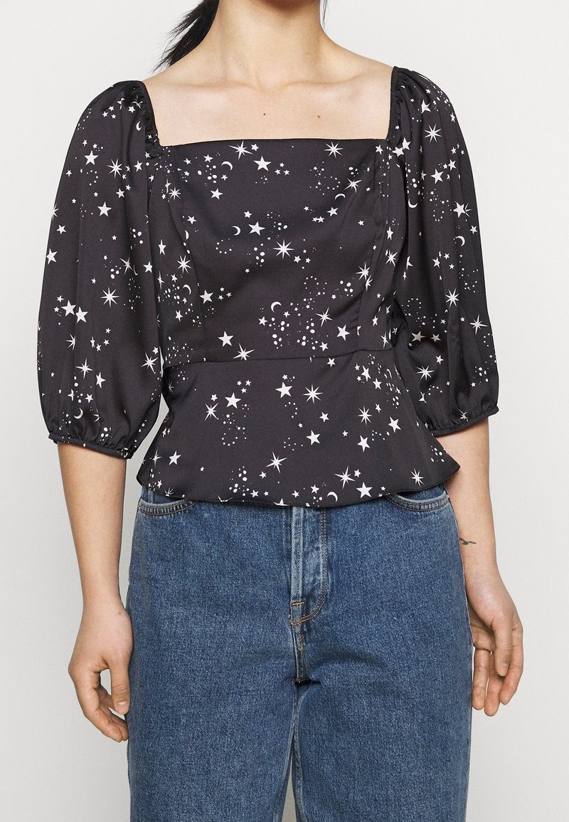 Blusa negra con escote cuadrado y mangas abullonadas, con patrones de estrellas y lunas blancas. Combinada con unos vaqueros de mezclilla azul de cintura alta.