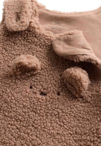 Brunt fleeceplagg med en texturerad, fluffig yta. Har björnöron, svarta broderade ögon och en mjuk beige krage. Tillverkat i Indien.