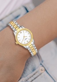 Orologio in acciaio inossidabile argento e oro con quadrante bianco, indicatori delle ore gialli e lunetta incisa, indossato su un polso contro un tessuto in denim.
