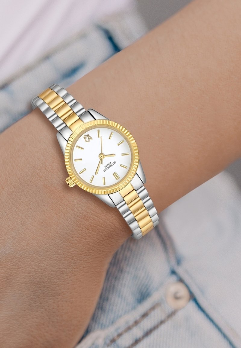 Orologio in acciaio inossidabile argento e oro con quadrante bianco, indicatori delle ore gialli e lunetta incisa, indossato su un polso contro un tessuto in denim.